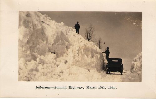 Jefferson-Summit Highway, March 15th, 1931. A man stands on top of a snow bank. Another man climbs onto his car to get on that snow bank.