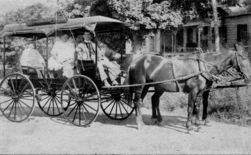 Two horses pulling a carriage of at least three people in the back. 