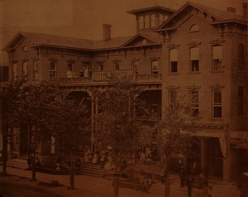 Large three story hotel with pillars, a balcony, a sign reading "Hotel Augustan" and several people both on the balcony and in front of the hotel.