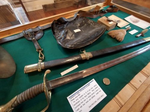 Part of a case showing a sword, a scabbard, a sword belt, a haversack, two brushes, a small bible, an identity disc, and two photographs.